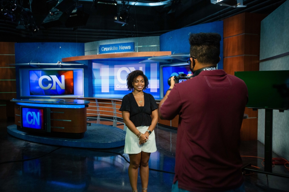 A college student smiles for a headshot with a news desk set behind her